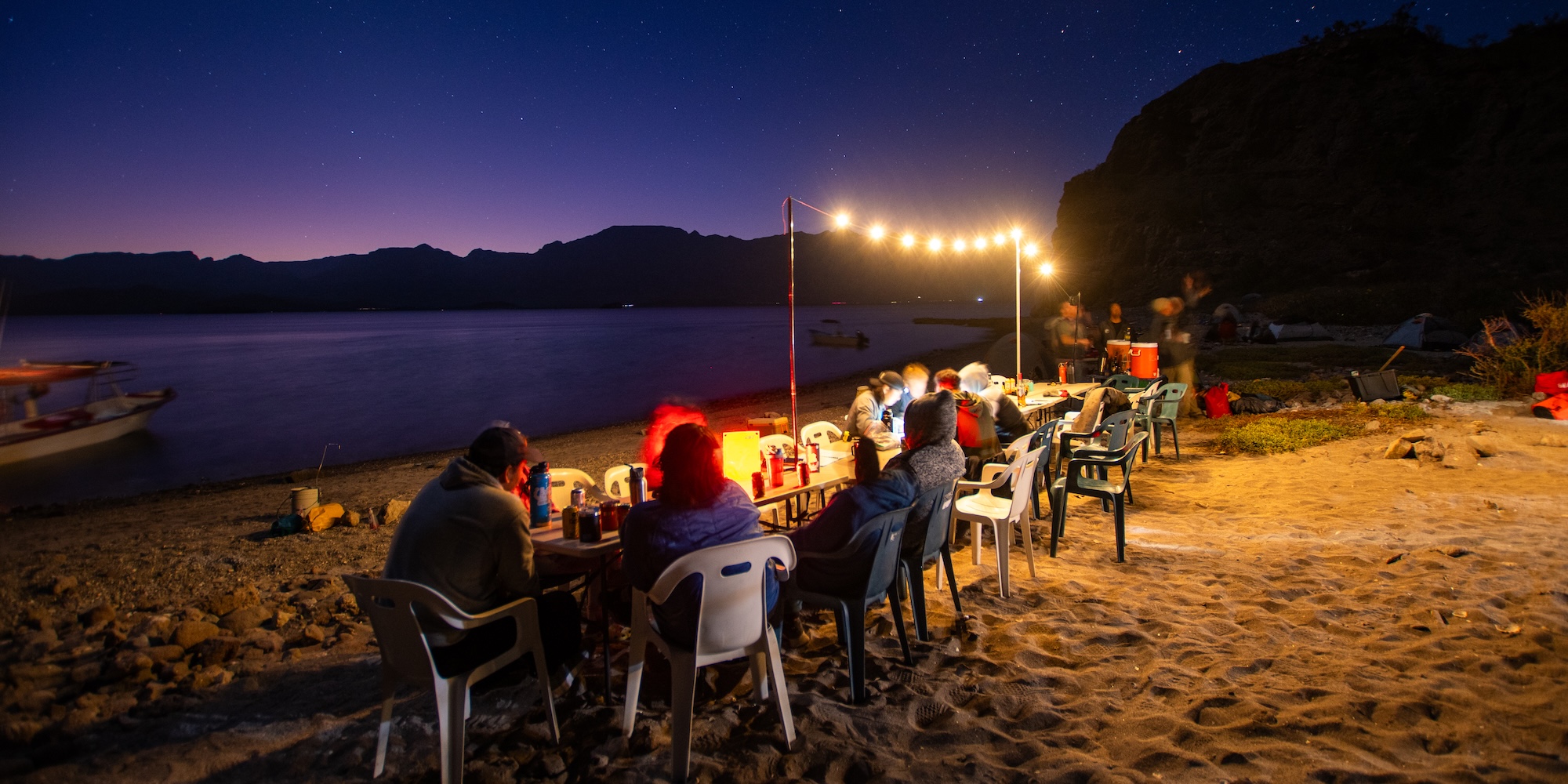 Travelers gathered around a glowing dinner table on a Baja beach at sunset, sharing stories after a day of sea kayaking under a starlit sky.