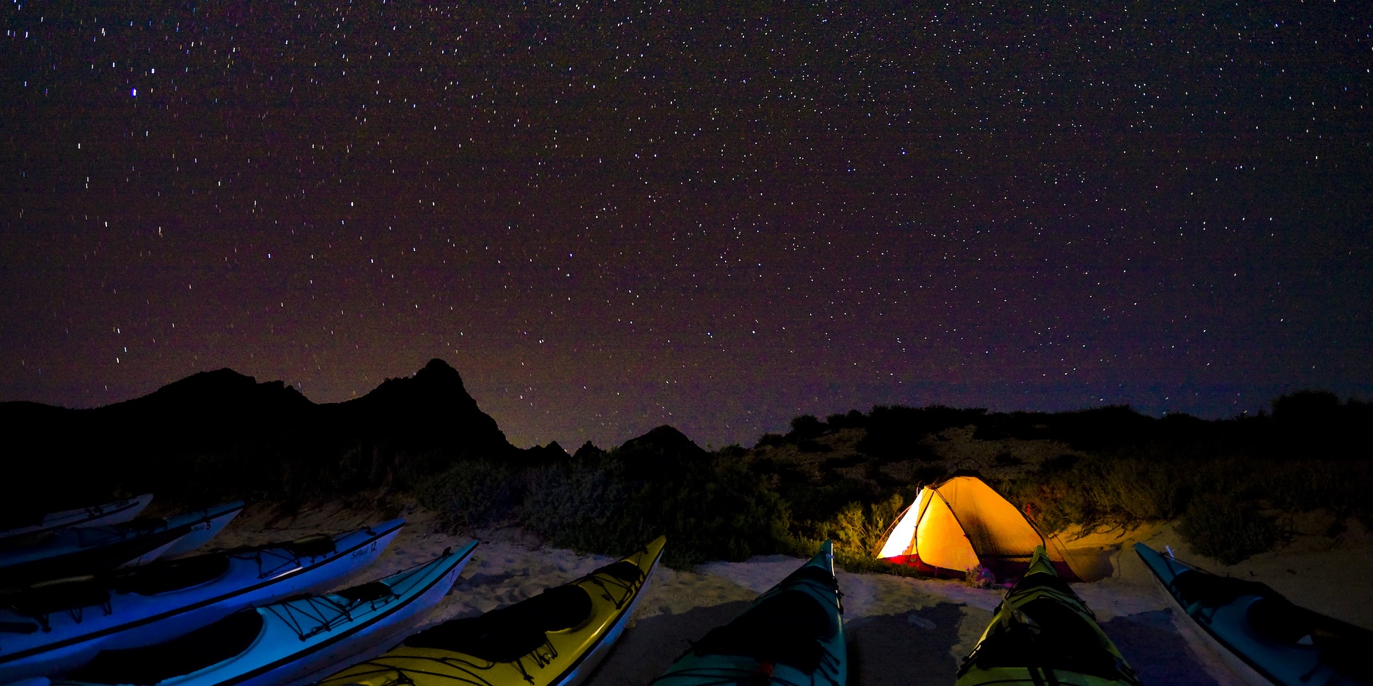 Kayaks lined up on a quiet beach beneath a blanket of stars, with a glowing tent nestled among Baja’s dunes at night.