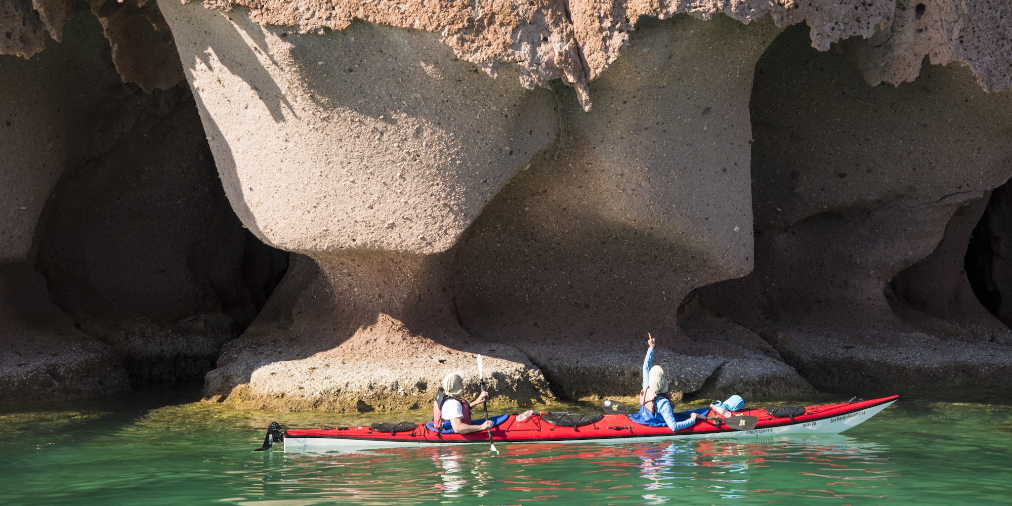 Two sea kayakers paddling beside dramatic rock formations in the turquoise waters of La Paz, Baja Mexico, exploring hidden coves and sea caves.